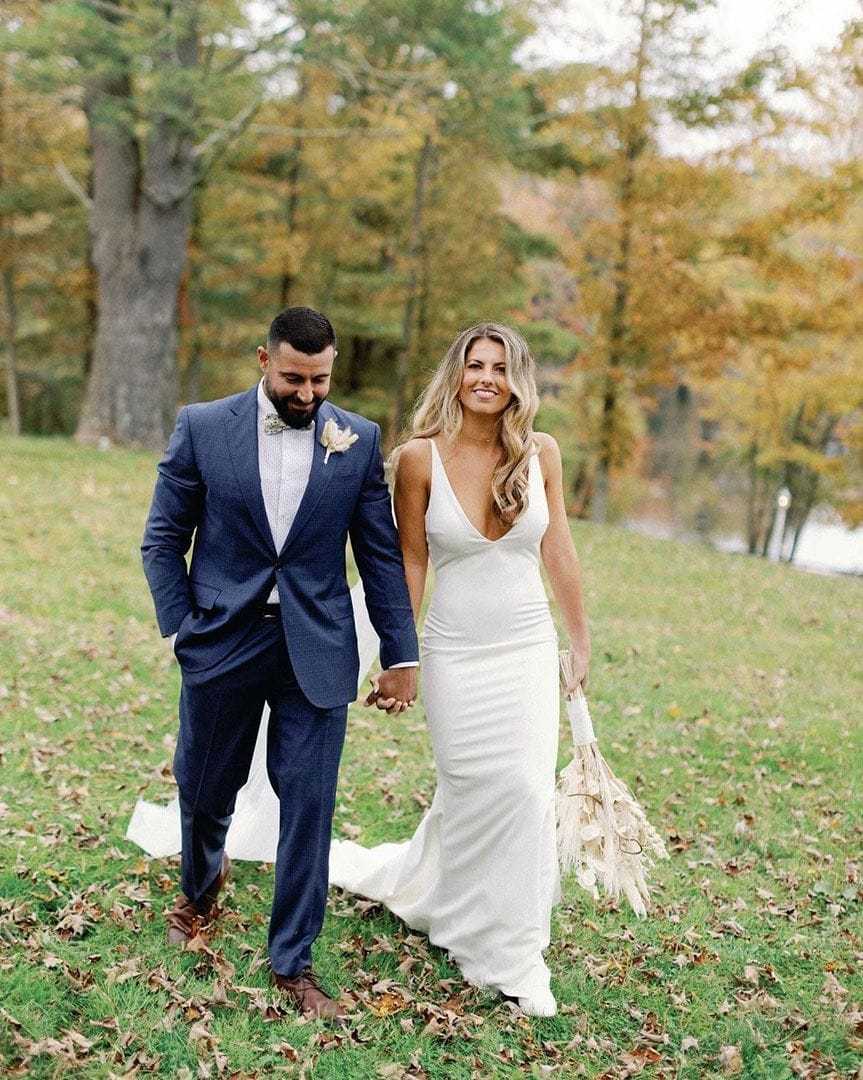 Bride in white dress and groom in blue suit walk hand in hand on a grassy path.
