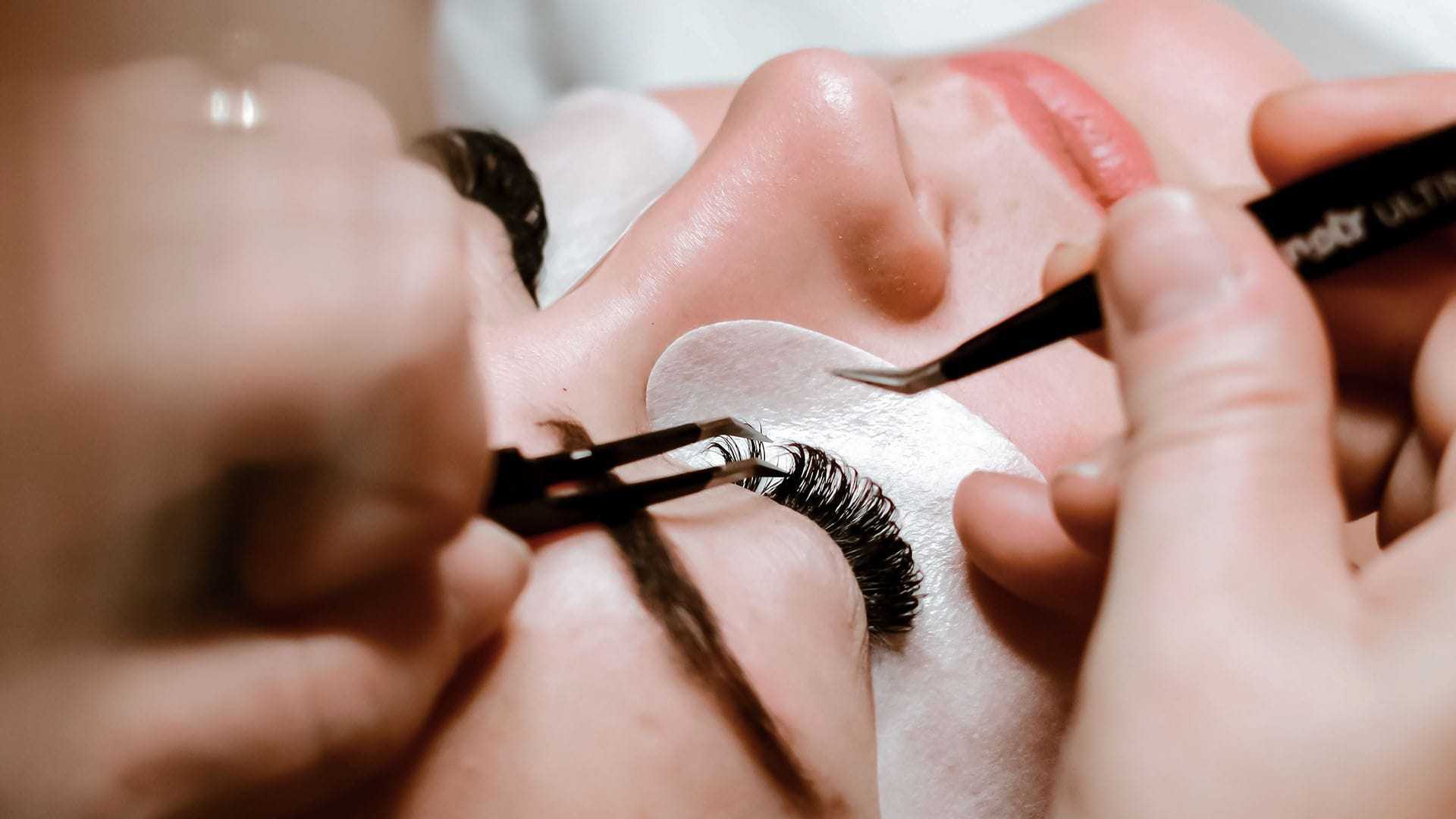 Applying eyelash extensions with tweezers, close-up of eyes and hands.